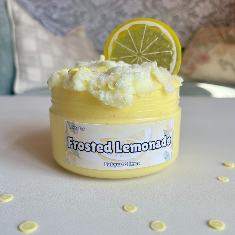 Peut inclure: Une slime citron glac&eacute;e jaune avec une tranche de citron sur le dessus. La slime est dans un pot transparent avec une &eacute;tiquette blanche qui dit "Frosted Lemonade" et "Babycat Slimes".