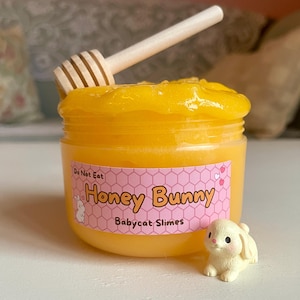 Könnte beinhalten: Ein Glas gelber Schleim mit einem hölzernen Honiglöffel. Das Glas hat ein rosa Etikett mit den Worten "Honey Bunny" und einer Hasengrafik. Eine kleine Hasenfigur sitzt neben dem Glas. Der Text "Do Not Eat" steht ebenfalls auf dem Etikett.