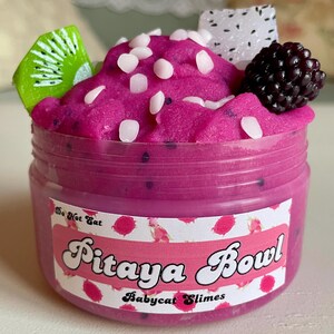 A round transparent container is full of magenta slime with tiny black seeds, resembling a pitaya dragonfruit bowl. on top are beeswax "coconut flakes" and fruit slice charms.