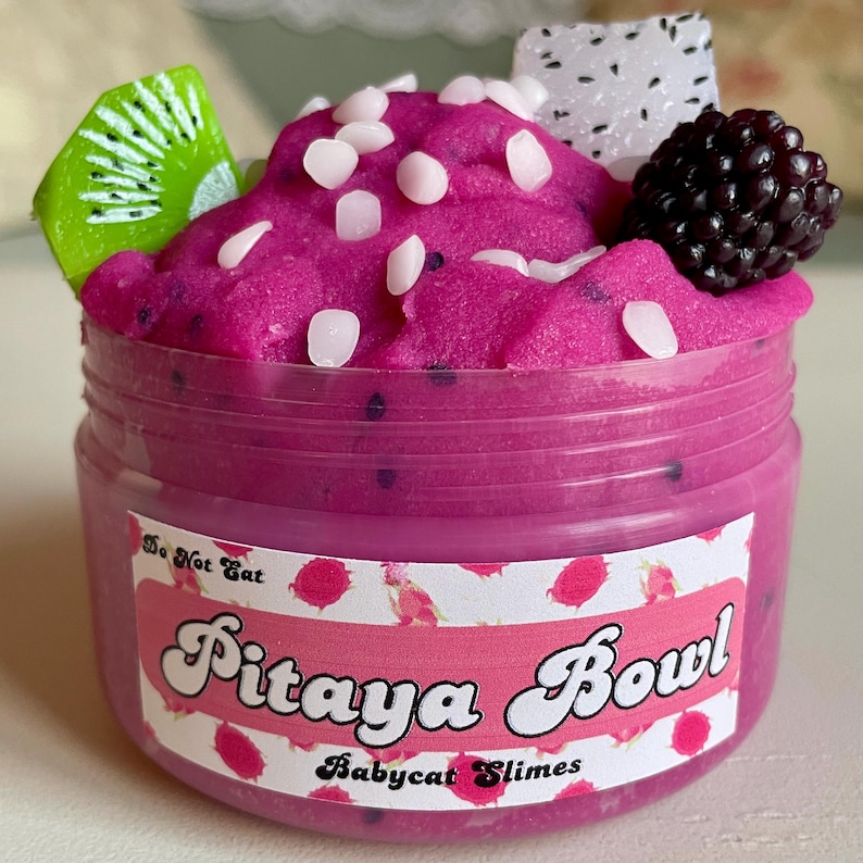 A round transparent container is full of magenta slime with tiny black seeds, resembling a pitaya dragonfruit bowl. on top are beeswax "coconut flakes" and fruit slice charms.