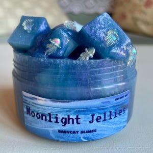 A clear, round plastic container is overflowing with blue sparkly jelly cube slime. The slime is a clear coated slime colorshifting from blue to purple, and has big squishable jelly cubes. It also has tiny opalescent jellyfish charms.