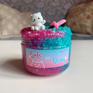 May include: A jar of colorful slime with a small cat figurine on top. The slime is a mix of pink and blue with glitter. The jar has a label that says "Cute but Stabby" with a cat illustration.