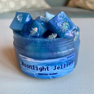 A clear, round plastic container is overflowing with blue sparkly jelly cube slime. The slime is a clear coated slime colorshifting from blue to purple, and has big squishable jelly cubes. It also has tiny opalescent jellyfish charms.