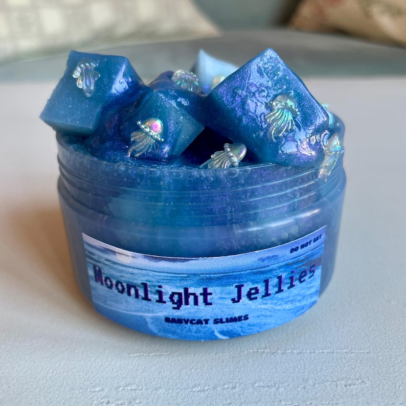A clear, round plastic container is overflowing with blue sparkly jelly cube slime. The slime is a clear coated slime colorshifting from blue to purple, and has big squishable jelly cubes. It also has tiny opalescent jellyfish charms.