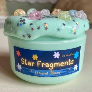 以下が含まれることがあります： Babycat Slimesの「Star Fragments」スライムが入った、明るい緑色の容器。スライムは、キラキラとカラフルな星型の紙吹雪、様々な宝石のような装飾で覆われています。容器のラベルには「Do Not Eat」と書かれています。