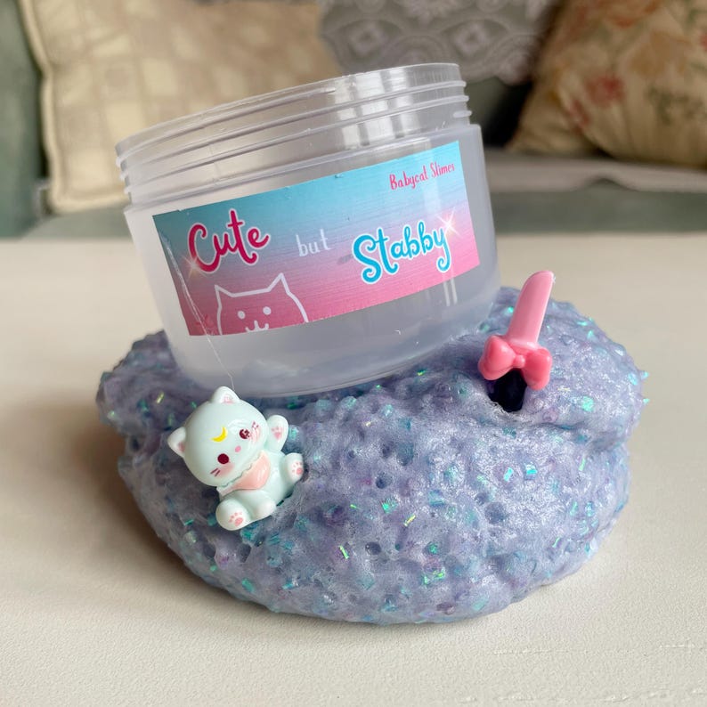 May include: A clear plastic container with the text "Cute but Stabby" on a label, resting on a bed of purple slime with glitter. A small pink bow and a light blue cat figurine are embedded in the slime.