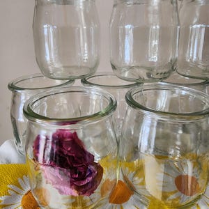 May include: A stack of clear glass jars, some with a dried pink rose inside. The jars are stacked on a yellow and white floral tablecloth.