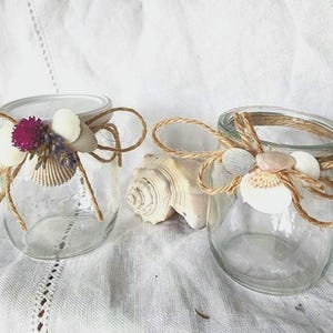 Könnte beinhalten: Zwei klare Gläser, verziert mit Muscheln, Bindfäden und kleinen Blumen. Ein Glas hat einen Deckel. Die Gläser sind mit weißen, rosa und lila Akzenten verziert. Die Gläser sind ein dekorativer Gegenstand.