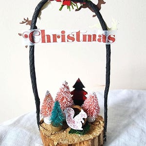 May include: A Christmas-themed decorative piece featuring a miniature forest scene. The scene includes small, colorful trees, a squirrel, and the word "Christmas" on a banner. The piece is set on a wooden base and framed by a black handle.
