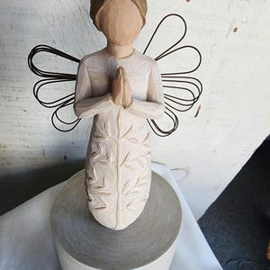 May include: A wooden angel figurine with wire wings, standing on a round base. The angel is carved with a floral design and has its hands clasped in prayer.
