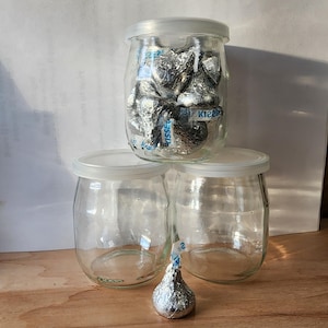 May include: Three clear glass jars with lids, stacked on top of each other. The top jar is filled with silver foil-wrapped chocolate kisses. A single silver foil-wrapped chocolate kiss is on the table in front of the jars.