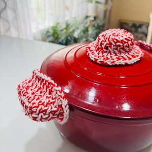 Puede incluir: Olla de hierro fundido esmaltada en rojo con agarraderas y tapa de ganchillo blanco y rojo.