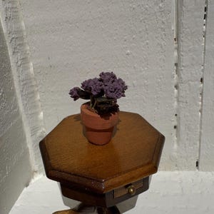 May include: A miniature potted plant with purple flowers sits on a small, hexagonal wooden table. The table has a drawer with a brass knob. The plant pot is terracotta-colored, and the table has a dark brown finish.