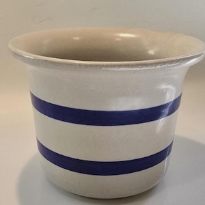 Puede incluir: Una maceta de cerámica con base color crema y dos franjas azules horizontales. La maceta tiene un borde ancho, ligeramente ensanchado y un cuerpo redondeado. El diseño es simple y clásico, adecuado para plantas y decoración.