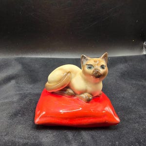 May include: A ceramic Siamese cat figurine resting on a bright red pillow. The cat has tan fur with dark brown markings on its face, ears, and paws. The cat's eyes are blue. The pillow is rectangular and glossy.