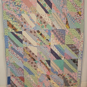 May include: A colorful patchwork quilt with a variety of patterned fabrics in shades of blue, pink, green, and white. The quilt features diagonal stripes and a floral border. The quilt is likely handmade and has a vintage aesthetic.