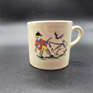 May include: Cream ceramic mug with a handle. The mug has a colourful illustration of a person in a red coat and yellow trousers, holding a rifle and walking a dog on a lead. The dog is grey and white.