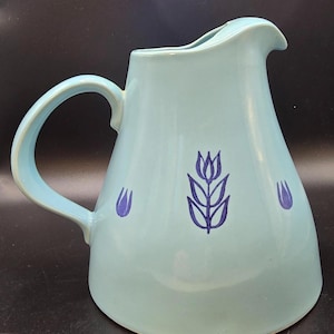 Può includere: Una brocca in ceramica azzurra con un manico curvo e un beccuccio. La brocca presenta un motivo floreale blu scuro e due piccoli accenti floreali. La brocca è un oggetto decorativo.