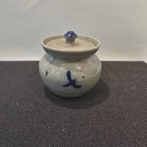 May include: A small, round ceramic jar with a lid. The jar is light gray with a blue design and a blue knob on the lid. The jar is sitting on a dark surface.
