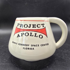 May include: Cream-colored ceramic mug with a rounded base and a large handle. The mug features the text "PROJECT APOLLO" in black letters with a red accent, along with "JOHN F. KENNEDY SPACE CENTER FLORIDA".