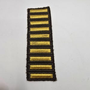 Könnte beinhalten: Ein rechteckiger Aufnäher mit acht horizontalen goldenen Balken, die auf einem dunkelbraunen Hintergrund gestickt sind. Die Ränder des Aufnähers sind ausgefranst. Die goldenen Balken haben eine strukturierte Optik, was auf ein militärisches Abzeichen hindeutet.