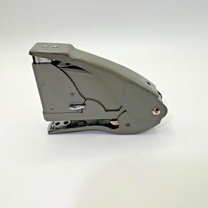 May include: A gray metal stapler with a curved design. The stapler has a silver metal base and a lever for operation. The body of the stapler is a muted gray color, and it sits on a white surface.