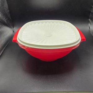 以下が含まれることがあります： 白い蓋が付いた赤いプラスチック製の容器。容器は角が丸く、赤いハンドルが2つ付いた四角い形をしています。蓋は白く、扇のような模様が施されています。食品の保存用です。