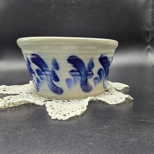 May include: A handmade ceramic bowl with a light beige exterior and blue abstract brushstrokes. The bowl sits on a white, crocheted doily. The bowl's design features a wide, open top and a slightly tapered base.