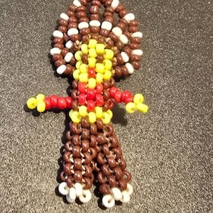 May include: A handcrafted beaded doll featuring a brown body, a head decorated with brown and white beads, and yellow and red details. The doll's arms and feet are also detailed with yellow and white beads. The doll is made of small, round beads.