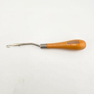 May include: A latch hook tool with a smooth, light brown wooden handle and a silver-colored metal shaft. The handle features the blue printed text readicut MADE IN ENGLAND. The metal hook end is bent at an angle for crafting purposes.