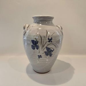 May include: A ceramic vase with a light gray, speckled glaze and a rounded body. The vase features two small side handles and a hand-painted design of dark blue flowers with green stems and leaves centered on the front of the vessel.
