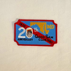Könnte beinhalten: Ein rechteckiger Aufnäher mit blauem Hintergrund und rotem Rand. Der Aufnäher zeigt den Text "20th Anniversary" und "Good Sam Club" mit einer Cartoonfigur und der Umriss des Bundesstaates Maryland. Die Jahre 1970 und 1990 sind ebenfalls abgebildet.