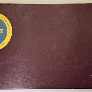May include: A vintage Scrabble game box with a dark burgundy exterior. A circular sticker on the box features the word "SCRABBLE" in yellow and blue, with additional text indicating the manufacturer and patent information.