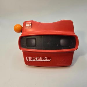 Könnte beinhalten: Ein roter View-Master-Betrachter mit schwarzem Sichtbereich und orangefarbenem Knopf. Das View-Master-Logo ist weiß, und das GAF-Logo befindet sich oben. Ein klassisches Spielzeug zum Betrachten von 3D-Bildern.