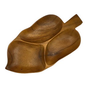 Pode incluir: Uma bandeja de servir de madeira em forma de folha, com três compartimentos. A bandeja é feita de madeira castanha clara e tem uma superfície lisa. Os compartimentos ovais são projetados para lanches ou itens pequenos.