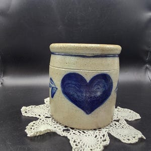 Puede incluir: Un tarro de gres con un diseño de corazón azul. El recipiente cilíndrico tiene un color base beige claro con un corazón azul oscuro pintado en la parte delantera. El borde y los detalles también son azules. El tarro está sobre un tapete de encaje blanco.