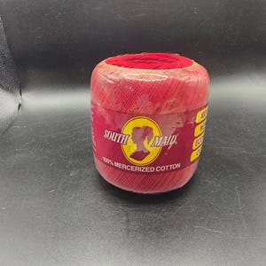 May include: A cylindrical spool of red thread, wrapped in clear plastic. The label features the text "South Maid" in a stylized font, with a silhouette of a woman's head. The label also states "100% Mercerized Cotton".