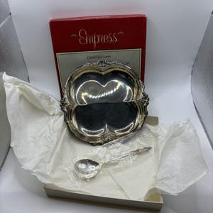 May include: A silver-plated clover-shaped dish with ornate edges and a matching silver-plated spoon in a plastic sleeve. The set is presented in a red and white box labeled Empress International Deepsilver, resting on white tissue paper.