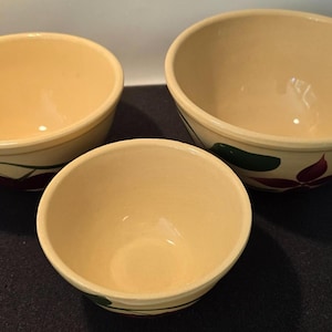 Puede incluir: Tres cuencos de cerámica color crema de diferentes tamaños. Cada cuenco presenta un diseño floral con detalles en rojo y verde. Los cuencos están dispuestos sobre una superficie oscura, mostrando su forma y detalles decorativos.