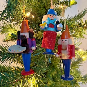 May include: Three wooden Christmas tree ornaments. One ornament is a painter with a palette, another is a figure holding a bird and a cup, and the third is a figure with a red coat. All have blue legs and orange hats.