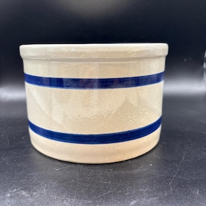 May include: A cylindrical stoneware crock with a beige background and two horizontal blue stripes. The rim and base are also beige. This vintage kitchenware item is likely used for food storage or decoration.
