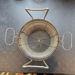 May include: A vintage, round metal fryer basket with a spiral wire interior. The basket has two handles on the sides and a larger handle on top. The metal has a slightly aged appearance.
