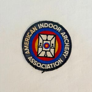 May include: Circular patch with the text "AMERICAN INDOOR ARCHERY ASSOCIATION" in white lettering on a black background. The patch features a red and blue bullseye design with a central square containing the letters "AOA".