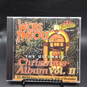 May include: A compact disc case featuring a cover with a pine tree background, a yellow and orange jukebox illustration, and text reading WCBS FM 101.1, The Ultimate Christmas Album Vol. II, Collectables COL-2512, and 25 MORE Great Songs for the 25th of December.