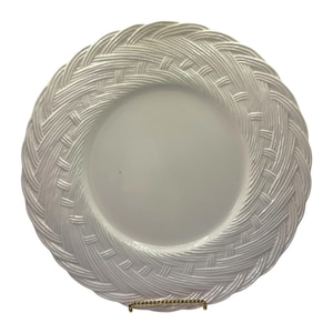 May include: White ceramic plate featuring a woven basket weave pattern around the edge. The centre of the plate is plain. The plate is round and is displayed on a gold-coloured stand.