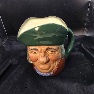 May include: A ceramic mug shaped like a man's head, featuring a green hat with a cream-colored brim. The mug has a handle and is painted with realistic facial features, including a red coat and blue patterned scarf.