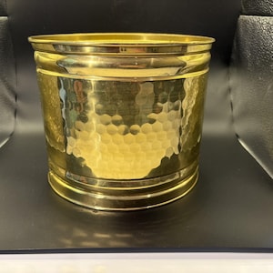May include: A cylindrical brass planter featuring a polished finish and a distinct hammered texture across its exterior surface. The vessel has a smooth, reinforced rim at the top and a matching base, showcasing a reflective metallic gold tone.