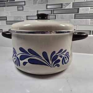 Op de afbeelding: Crèmekleurige emaille pot met een donkerbruin deksel en handvatten. De pot heeft een blauw bloemmotief rond de basis. Keukengerei.