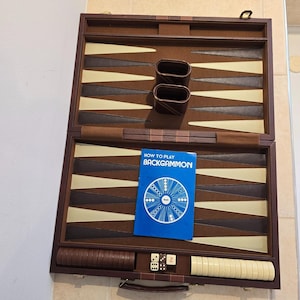 Puede incluir: Juego de backgammon abierto, con exterior de cuero marrón y superficie de juego con triángulos marrones y beige alternados. Incluye dados, fichas y un folleto azul titulado "HOW TO PLAY BACKGAMMON".