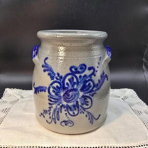 Puede incluir: Un tarro de gres gris con diseños florales azules. El tarro de cerámica tiene dos asas azules y un patrón floral azul en la parte delantera. El tarro está sobre un tapete de encaje blanco.